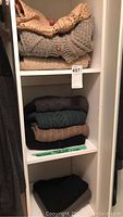 Shelf with folded sweaters including a gray cable knit cardigan, and other neutral-toned sweaters in brown, green, and black folded underneath and on bottom shelf