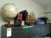 Wide view showing spinning globe on wooden base, two desk clocks, model ship, photo albums and various items on top of dark cabinet