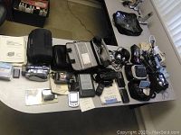Overall lot view showing a variety of electronic devices and accessories including cameras, recorders, label maker, GPS, Kindle, cables and media storage spread on a table