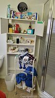 Overview of white wall cabinet with open doors containing soaps, personal care items, first aid supplies, small mirror on wheelchair, and wire shelf beneath holding hairdryers and neck bags.