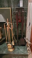 Two fireplace tool sets placed side by side on a green marble hearth: a polished brass-plated set on left with arched stand and hanging tools, and a tall black metal set with a rectangular design on right.