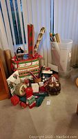 Photo of various Christmas wrapping paper rolls, pine cones, basket, tins, candles, and cards on carpeted floor in front of vertical blinds.