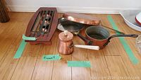 All items grouped on floor showing 2 copper frying pans, copper saucepan, teapot with wooden handle, and wooden wrought iron food warmer with tealight holders.