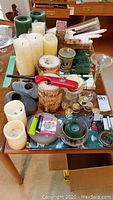 Wide view of assorted candles and candle holders arranged on a wooden table, including pillar candles, tapered candles, a green candle in a glass bowl with decorative stones, a red pocket knife, and various candle holders.