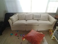 White fabric couch with five matching cushions placed in front of large windows. Has subtle texture and curved armrests. Slight stain visible on bottom right side as provided by seller. A red cushion and some children's toys are visible in the foreground.