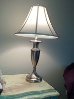End table lamp with pewter base lit up on a distressed wooden table with greenish tint.
