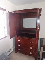 Open upper cabinet of dark brown wooden armoire showing interior space with back panel and cable opening, and three storage drawers below