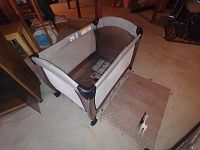 A beige and brown portable pack and play playard with mesh sides and a foldable padded floor mat lying flat on the ground in a room with wooden furniture.