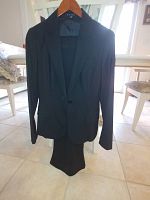 Photo of a black ladies pants suit by Theory, hanging on a wooden hanger in a light-filled room, showing classic tailored blazer and matching pants.