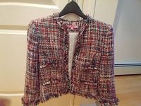 Front view of a multi-color woven ladies jacket on a black hanger. The jacket is cropped with fringe trim detailing on the collar, cuffs, pockets, and hem. Two front flap pockets.
