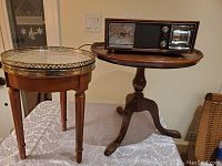 Photo shows two small wooden tables side by side, one with a marble top and metal rim, the other an oval wooden table with a pedestal base. The vintage radio with clock is placed on the oval table.