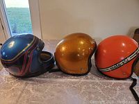 Photo showing 3 vintage helmets: blue Kawasaki full-face, gold glitter Innov open-face, and orange moto-ski open-face helmet.