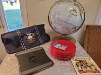 Large world globe, 8 Fiesta plates, navy blue tote bag with owl design, large black laptop case, cookbook and package of blue dye on table.