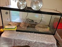 Front view of the large glass reptile aquarium tank showing inside with sand substrate, two reptile hides, water dish and plastic container with lamps on top.