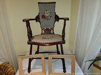 Front view of vintage armchair with carved wooden legs and armrests, needlepoint seat and backrest with floral design, two wooden frames in front.