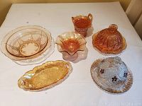 Full lot overview showing 2 amber carnival glass butter dishes, 1 basket pitcher, 2 candy dishes, and 1 bowl arranged on table.