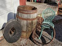 Photo showing large wooden barrel with metal rings, round barrel top with cut-out, decorative green metal chair, and extra metal hoops on the ground in front of a porch.