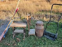 Wide view of lot outdoors on grass showing all items: old milk can, push lawn mower, stool, wooden tool and rod.