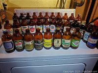 Front view of 32 vintage brown tubby beer bottles showing various brand labels arranged on a white surface.
