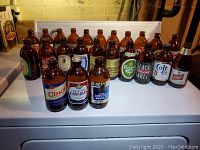 24 assorted older stubbie beer bottles displayed in rows showing a variety of labels and bottle caps, placed on white surface.
