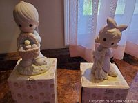 Both Precious Moments figurines standing on their original boxes on a table near a window with a curtain