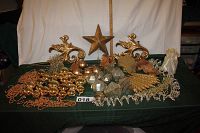 Overall display of gold ornaments, beads, star tree topper, ribbons, angel figurine, two bird ornaments, gold bells, and other assorted decorations on green cloth with white background and measuring tape.
