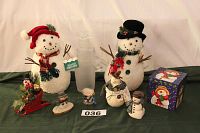 A lineup of seven snowman decorations including two large white snowmen with twig arms and hats, one tall frosted glass snowman, several smaller snowman figures, and a decorated snowman-themed box arranged on a green surface.