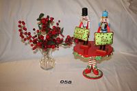 Photo of two nutcracker figurines labeled Naughty and Nice standing on a red and green ceramic cake stand with a figure base dressed in striped stockings and elf shoes, next to a small vase with grape and green leaf arrangement.