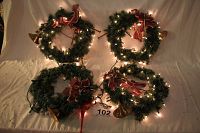 Four green Christmas wreaths laid flat showing brass horns, red ribbons, and string lights with three lit and one unlit.