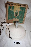 Photo shows the off-white speckled round base of the tree stand with attached electrical cord and chrome tree holder, placed in front of original cardboard box with visible wear and tape.