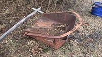 Overall view of the metal bucket scoop and wooden handle of a scythe lying on grass and dried leaves.