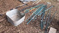 Galvanized tin water tub sitting on ground with a pile of green metal fence posts beside it outdoors on leaf-covered soil.