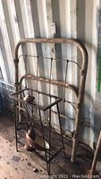 Rusty iron twin bed frame leaning against a wall with a metal rectangular stand in front containing lamp housing parts.