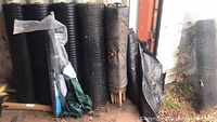 Overview showing five large rolls of black plastic-coated fencing, green shade cloth and wooden stakes at base