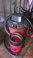 Two red Shop Vac wet/dry vacuums with attached hoses, one missing the top motor, in a workshop setting.