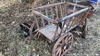 Front angle showing the wooden Greek market cart with spoked wheels and visible metal components, placed outdoors with dry grass and dirt ground.