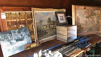 Wide view showing the six framed paintings leaning against a wall and a wooden organizer box on a wooden surface. Paintings depict landscapes, nature, and abstract scenes with wood frames showing wear.
