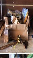 Box containing hand tools such as hammer, chisel, axe head, sledgehammer head with wooden handles and metal heads, along with bags and boxes of hardware parts