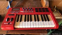 M-Audio Axiom 25 red keyboard controller on wooden bench, showing keys, pads and knobs.