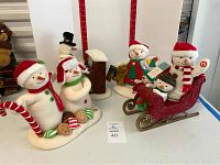 Five Hallmark plush snowmen with Christmas themes displayed on a flat surface, including two in red sleigh and one playing piano.