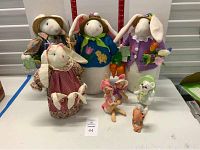 Overview of fifteen stuffed garden bunnies, mice, and deer dolls with different outfits and accessories arranged on table.