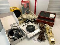 Bose headphones in original box with manual and earpiece covers degrading, portable Bose CD player, multiple white surge protectors, multi-plug extension cord on yellow reel, decorative remote for holiday lighting, and wooden compartment organizer box.