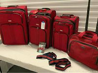 Four red Ricardo Elite luggage pieces displayed on white cloth surface; three upright suitcases of varying sizes and one smaller carry bag with handles and shoulder strap in front.