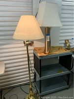 Two lamps in storage unit: floor lamp on the left with white tapered shade, metal stand; table lamp on the right with short cylindrical golden base and square white shade on a small black cart.