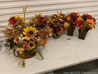Three pairs of fall floral artificial arrangements on a white table in front of a storage door. Arrangements include sunflowers and other autumn flowers in woven baskets and square containers.
