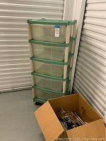 Full view of Rubbermaid five-drawer plastic storage unit on rolling cart and cardboard box with hangers beside it.