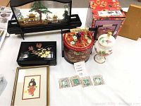 Overview of all Asian decorative items on table including jewelry box, framed art, music box under glass, porcelain vase, small ceramic tiles, and boxed items