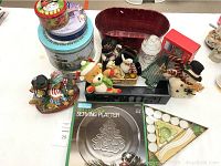 Photo showing Christmas tins, a rusty red planter, a clear glass jar, various Christmas figurines, and a boxed serving platter with tree design