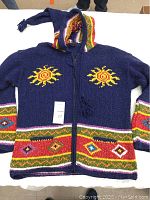 Front view of navy blue handmade Ecuadorian sweater with colorful sun and geometric patterns and attached hood with tassels, laid flat on table.