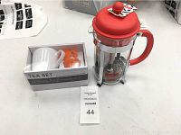 Photo showing the boxed Home Art white ceramic tea mug with orange silicone flower-shaped tea steeper next to a Bodum coffee press with red lid and handle on a white surface.
